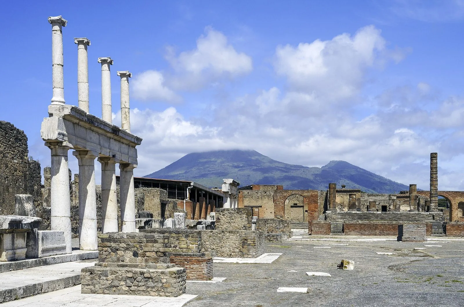 Il Foro di Pompei con le colonne del portico in primo piano, il Tempio di Giove sul fondo e il Vesuvio sotto un cielo con nuvole
